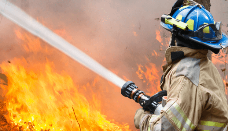 A firefighter sprays water on a wildfire, wearing full protective gear with smoke and flames in the background.