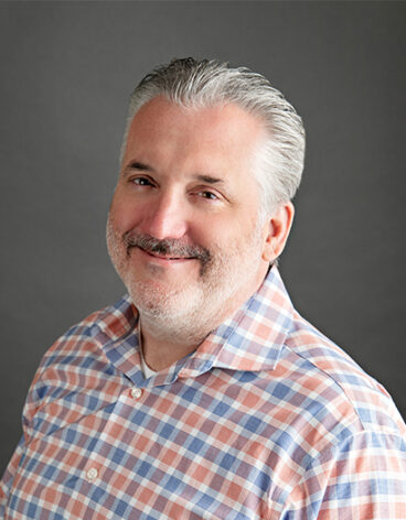 A person with grey hair and beard wearing a plaid shirt
