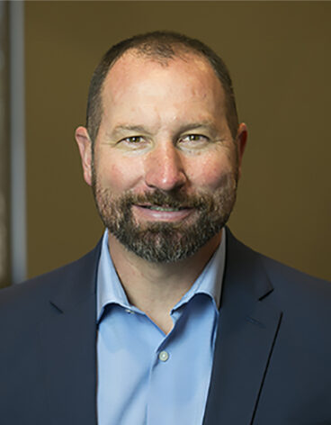 Man smiling in a blue suite with a blue shirt in front of a tan wall.