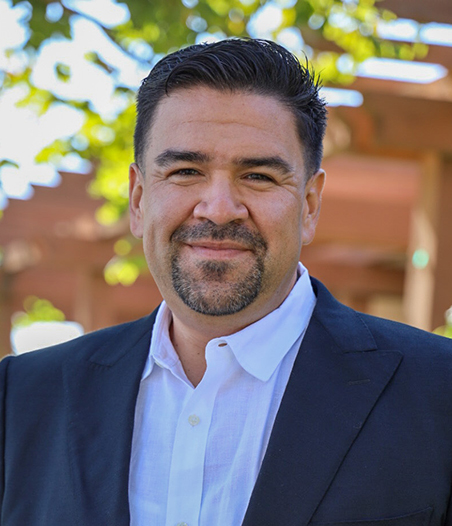 Person smiling in a blue suit and white shirt in front of a tree