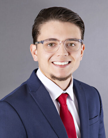 Man smiling with short brown hair, a red tie, and navy blazer.