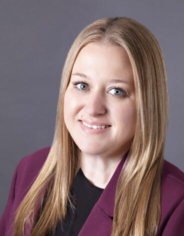 Woman smiling with dark blonde hair and a maroon blazer.