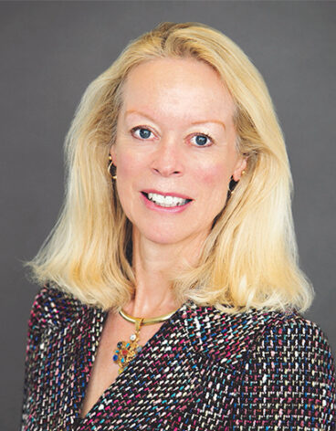 A person smiling wearing a pearl necklace and coral blazer.