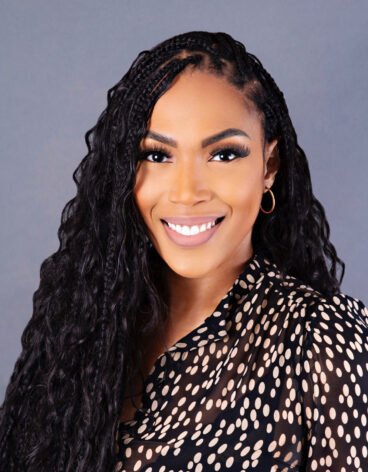 Woman smiling with long dark hair and a spotted blouse.