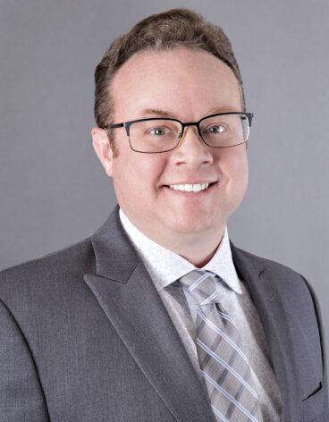 Man smiling with short light brown hair and a gray blazer and tie.