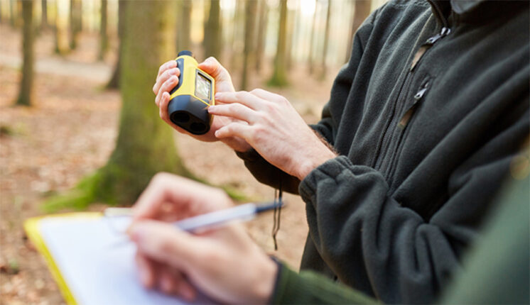 The hands of two individuals holding a clipboard and small pieces of equipment with trees in the background