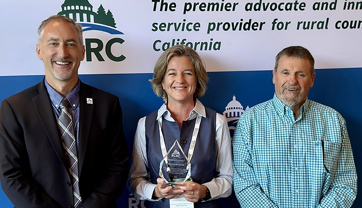 Three individuals smiling with the woman in the middle holding an award.