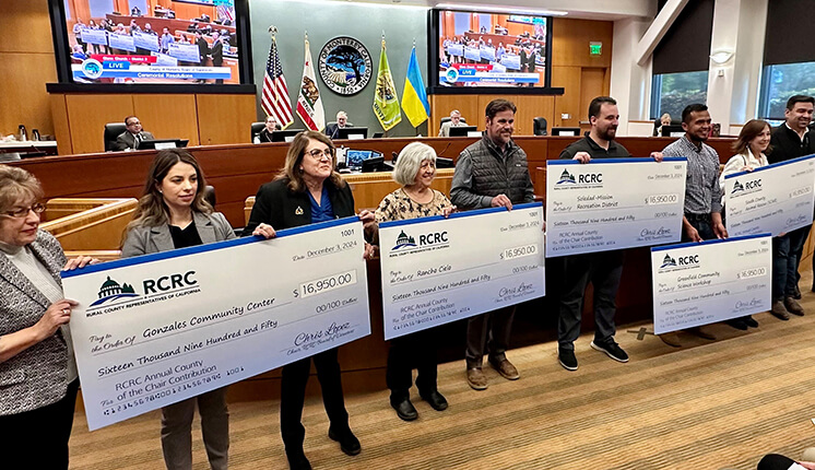 A group of people holding large checks in a county board room.
