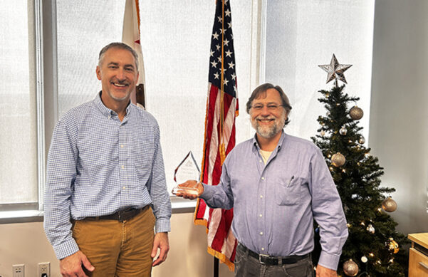 Two men standing in front of the U.S. flag smiling, one is holding an award.