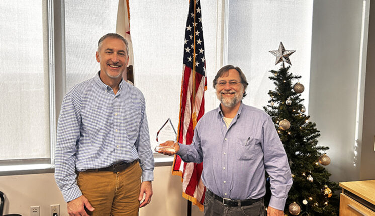 Two men standing in front of the U.S. flag smiling, one is holding an award.