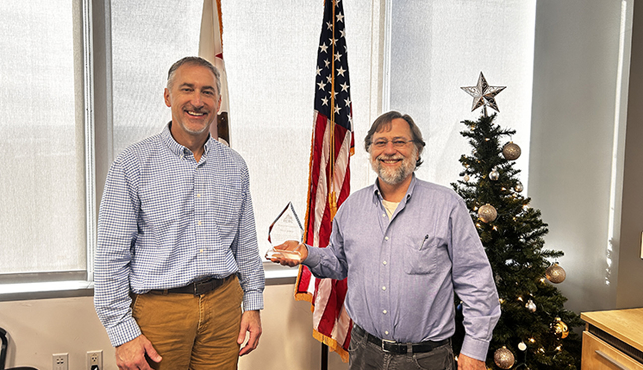 Two men standing in front of the U.S. flag smiling, one is holding an award.