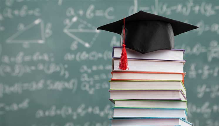 graduation cap stacked on books, chalkboard blurred in background