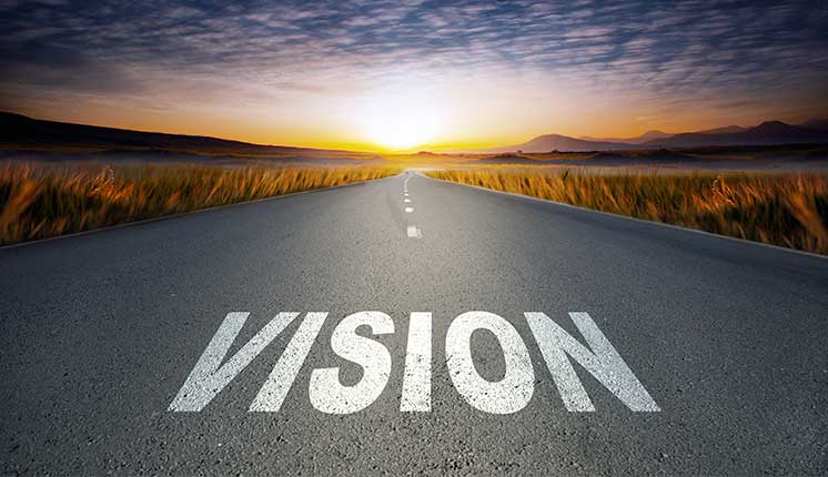 the word "Vision" on top of a road with fields beside and distant mountains awash in the light of the rising sun