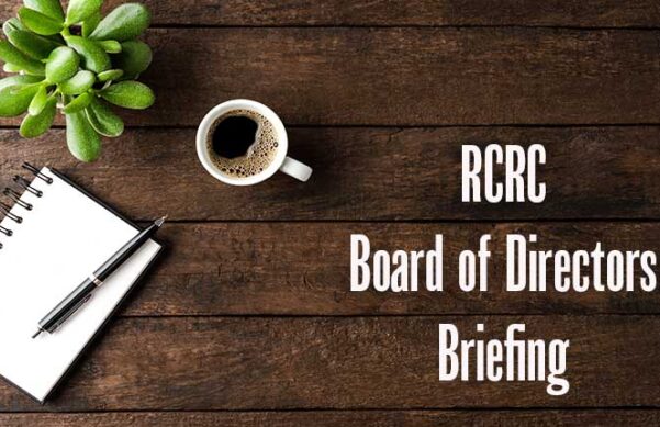 overhead view of coffee mug, notepad and pen, plant on wooden backdrop with the words RCRC Board of Directors Briefing
