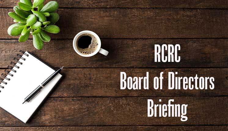 overhead view of coffee mug, notepad and pen, plant on wooden backdrop with the words RCRC Board of Directors Briefing